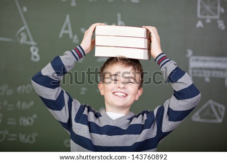 Happy male student carrying stacked books on head against chalkboard