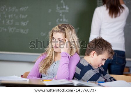 Bored students sitting at desk with teacher in background at classroom