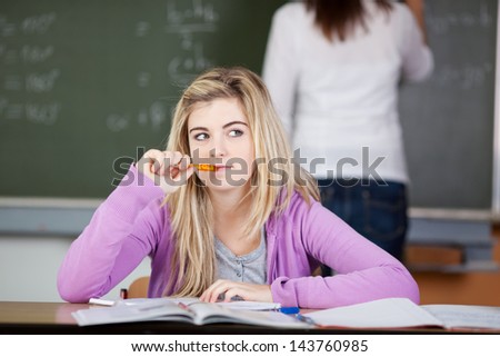 Thoughtful female student sitting at desk with teacher in background at classroom
