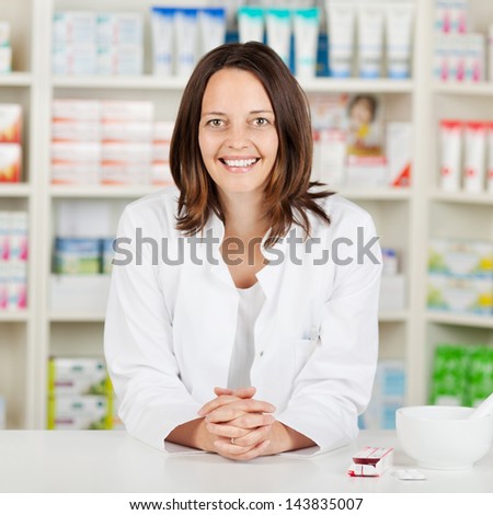 Portrait of mid adult female pharmacist with hands clasped leaning on pharmacy counter