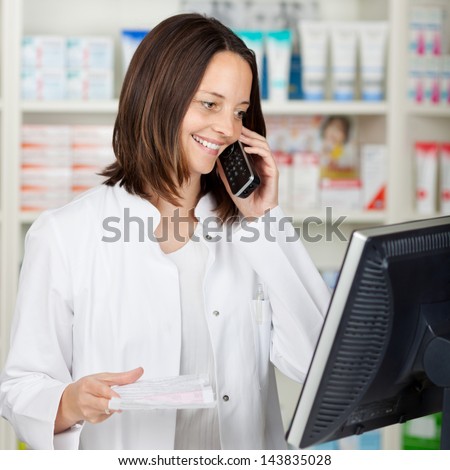 Mid adult female pharmacist using cordless phone while looking at computer in pharmacy
