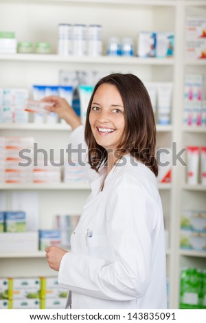 Portrait of happy female pharmacist taking medicine from shelves at pharmacy