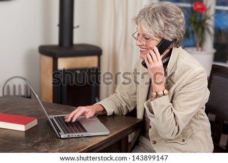 Side view of a smiling female pensioner using laptop and mobile phone at home