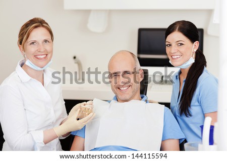 Dentist explaining teeth model to male patient at clinic while looking at camera