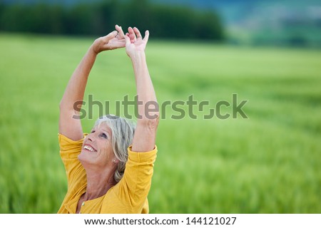 Happy Senior Woman With Arms Raised Looking Up While Standing On Grassy Field