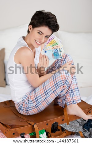 Full length portrait of happy young woman sitting on overloaded suitcase while holding money fan in bed