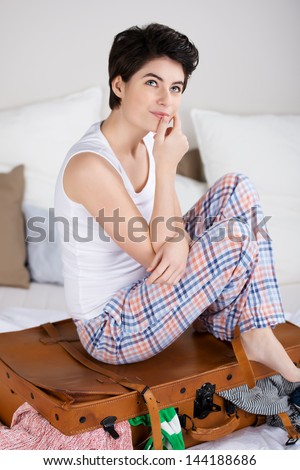 Beautiful young woman in her sleepwear sitting on her bed on top of her packed suitcase dreaming of a forthcoming trip