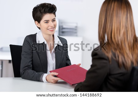 Happy young female candidate giving file to businesswoman at desk in office