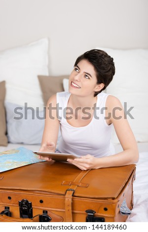 Beautiful young woman leaning on a large brown leather suitcase planning a vacation and journey on a tablet