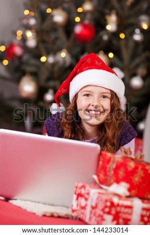 Beautiful young girl wearing a red Santa hat lying on the floor with her laptop computer in front of a decorated Christmas tree with sparkling lights surrounded by gifts