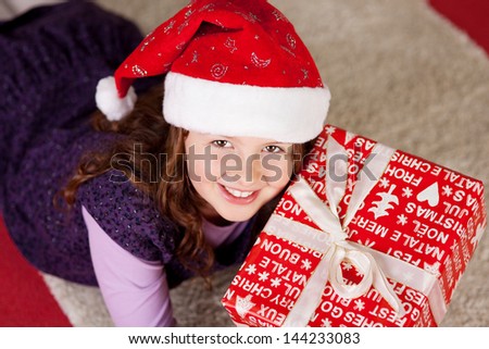 High angle view of a smiling young girl wearing a colorful red Santa hat lying on the floor with a large Christmas present looking up with a smile
