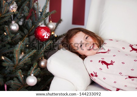 Cute smiling young girl lying on her back on a sofa alongside the Christmas tree waiting for the Christ Child draped in a red and white cloth with reindeer