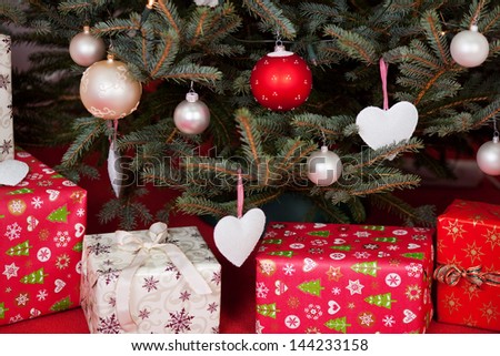 Gift boxes wrapped in decorative paper under the Christmas tree, with ornamental globes and heart shapes hanging