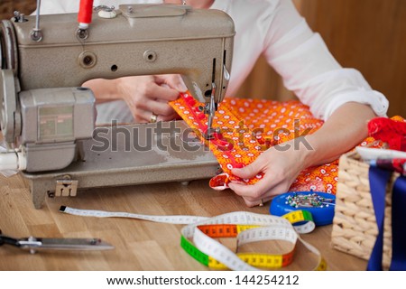 Seamstress with her sewing machine sewing a border onto a colorful orange item of clothing as she works at her table