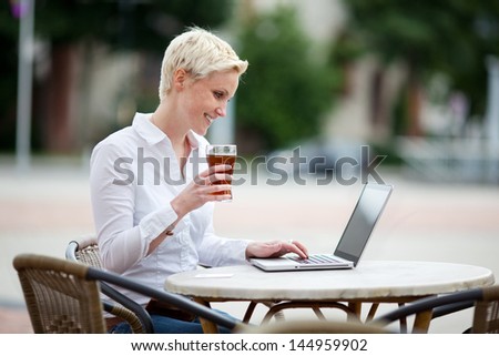 Happy young blond woman using a laptop at an open-air restaurant sitting outdoors at a table on the street holding a cold drink in her hand