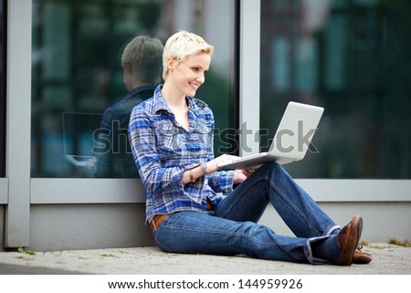 Student with laptop in the city leaning against a glass building