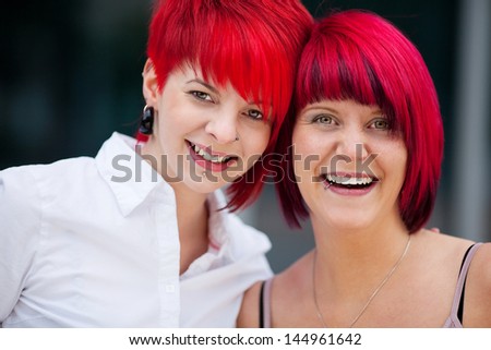 Two beautiful modern fashion conscious women with fiery dyed red hair and modern haircuts standing together smiling at the camera, head and shoulders portrait