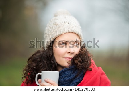 Closeup of beautiful young woman in winter clothes looking away while holding coffee mug in park