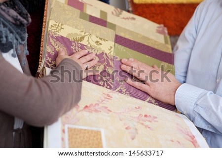 Woman looking at wallpaper and fabric swatches holding a sample book in her hands while discussing them with a male partner or salesman