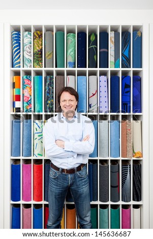 Motivated attractive young salesman standing with his arms folded in front of a colorful carpet display in an interior decorating shop