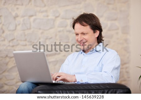 Smiling man sitting in an armchair working on a laptop at home against a painted stone wall with copyspace