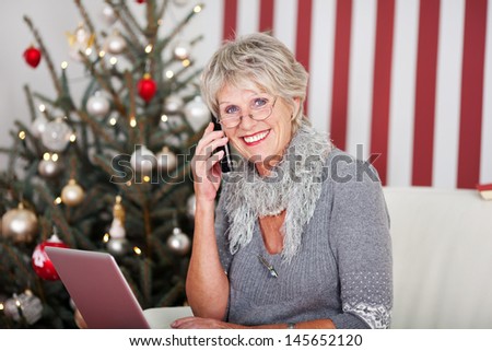 Senior woman chatting on the telephone at Christmas wishing all her friends a Joyful Christmas while sitting on a sofa in front of the Christmas tree