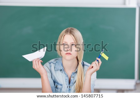 Thoughtful female student holding paperboat and flag against chalkboard in classroom