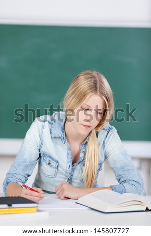 Young woman busy studying reading from a large textbook and taking notes on a notepad sitting in front of a class blackboard