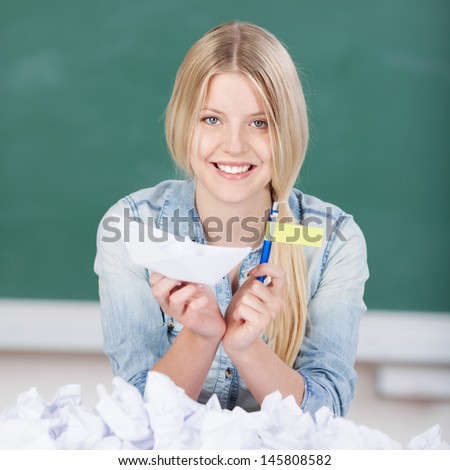 Portrait of young female student smiling while holding paperboat and flag with paperballs in foreground at classroom