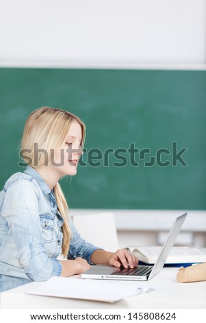 Attractive young college student working at her laptop sitting reading the screen and typing in front of a blank green blackboard