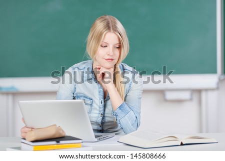 Young female student working with a laptop and textbook in the classroom in front of a blank green blackboard