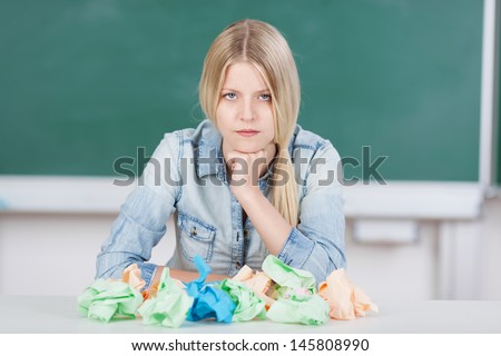 Pretty young blond student or teacher with writers block sitting in front of the blackboard in the classroom in front of a pile of crumpled paper with a despondent expression