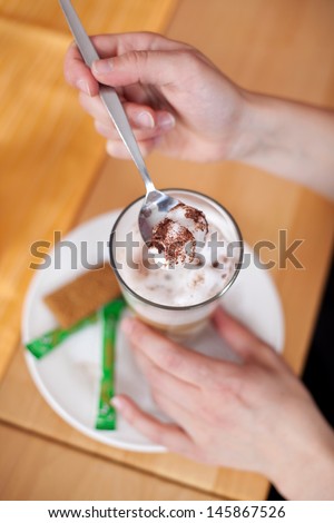 Closeup overhead view of a woman enjoying a latte macchiato coffee taking a spoonful of the frothy cream from the top of the glass