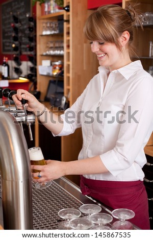 Pretty young waitress serving draft beer standing behind the bar counter dispensing it into a pint glass from a metal spigot