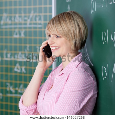 Beautiful young student standing in front of the blackboard chatting on her mobile in the classroom