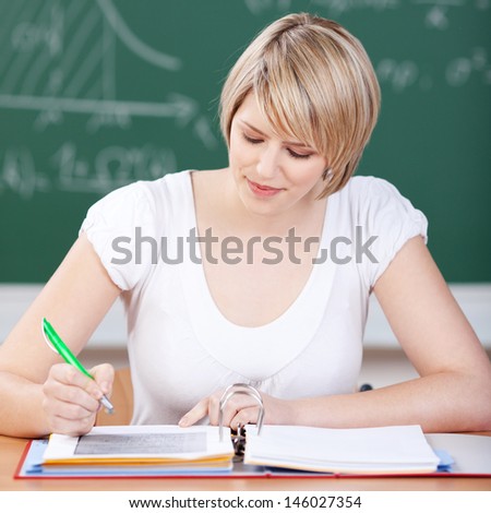 Attractive young female student sitting at a desk with an open binder of notes working in class in front of the blackboard