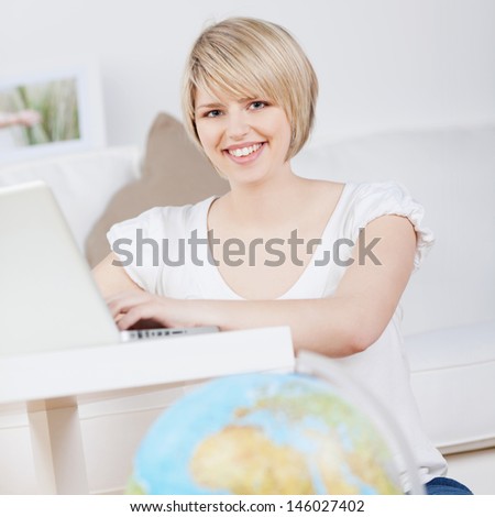 Smiling young woman sitting at her laptop computer booking her holiday reservations online with a world globe in the foreground, focus to the womans face
