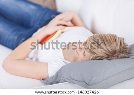Woman lying on her back on a sofa falling asleep while reading a book which is resting on her chest
