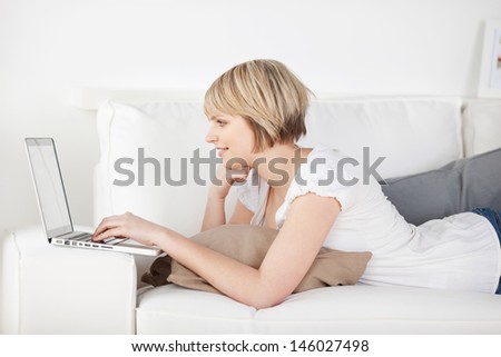 Profile view of a young woman working with a laptop on the sofa lying stretched out on her stomach with the computer balanced on the arm
