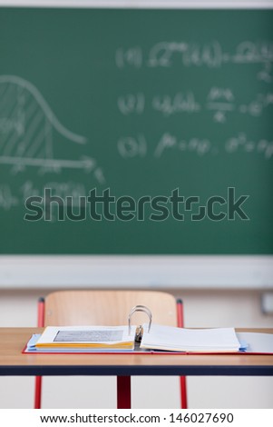 One vacant student desk in the classroom with an open binder containing class notes in front of a green blackboard