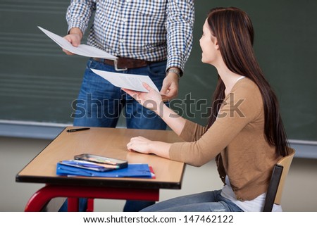 Midsection of male professor collecting answer sheet from young woman at desk in classroom