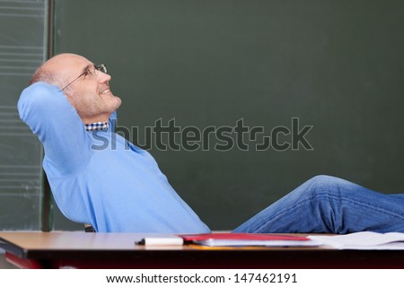 Relaxed thoughtful male teacher with hands behind head looking up at desk in classroom
