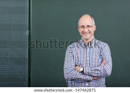 Portrait of confident male professor with arms crossed standing against chalkboard in classroom