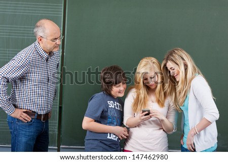 Angry teacher looking at students using mobilephone against chalkboard in classroom