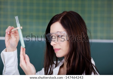 Young woman wearing plastic safety goggles checking on a chemical reaction in a test tube she is holding up while doing university research