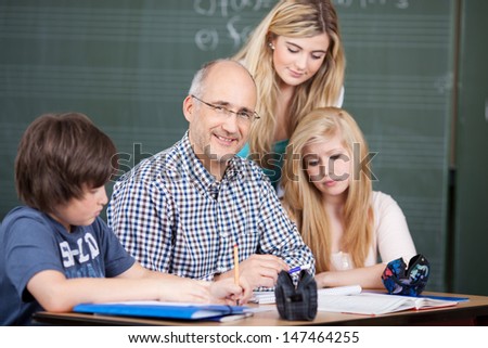 Friendly middle-aged male teacher sitting at a desk with class notes surrounded by his young pupils, two attractive teenage girls and a boy