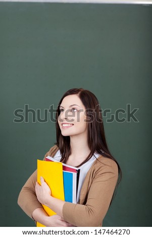Pretty young female student standing thinking clutching a colorful set of folders to her chest against a dark grey background with copyspace