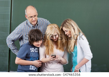 Schoolchildren playing with a mobile phone grouped together peering at the message on the screen with their male teacher looking over their shoulders trying to see what they are doing