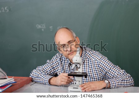 Middle-aged male teacher sitting at his desk in the classroom in front of the blackboard looking at a microscope