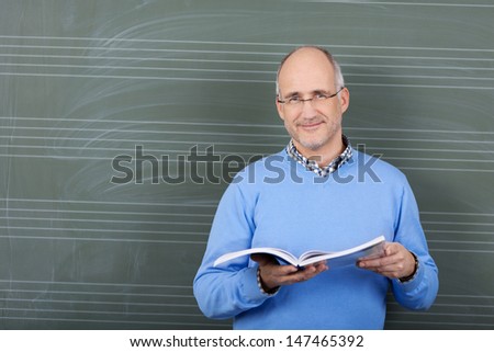 Smiling male teacher with a text book in his hands standing in front of the blackboard in the schoolroom with copyspace